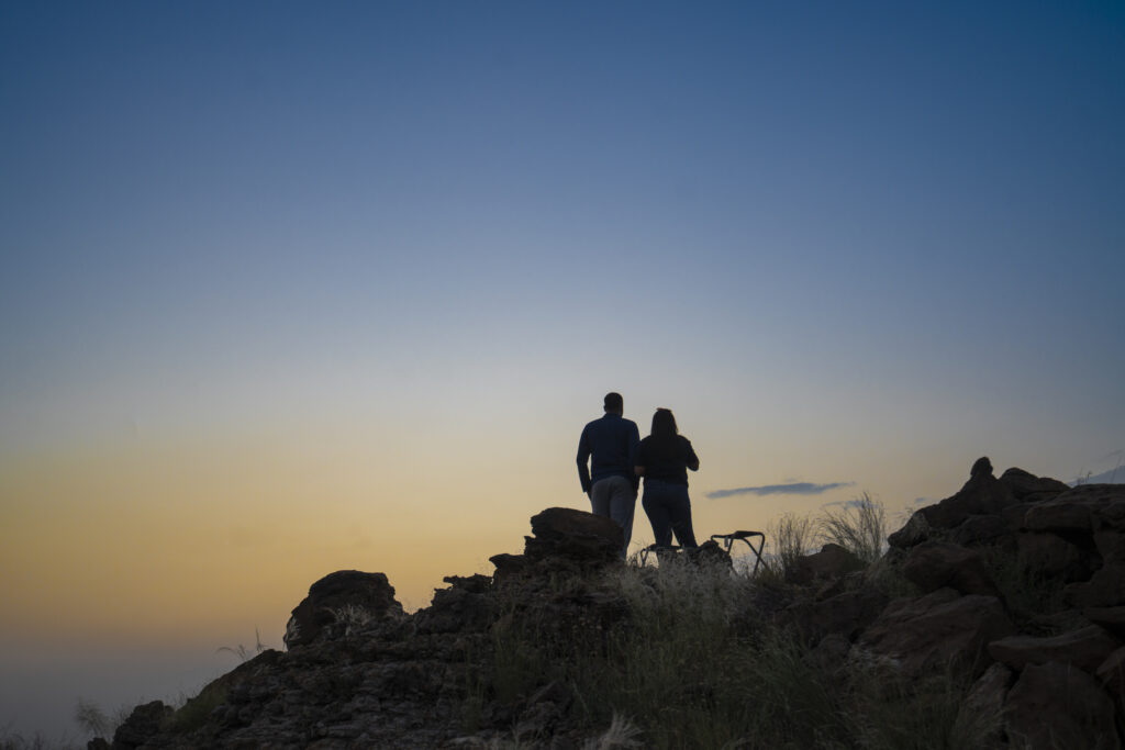 Couple standing on rocky desert hill, watching peaceful sunset sky together.