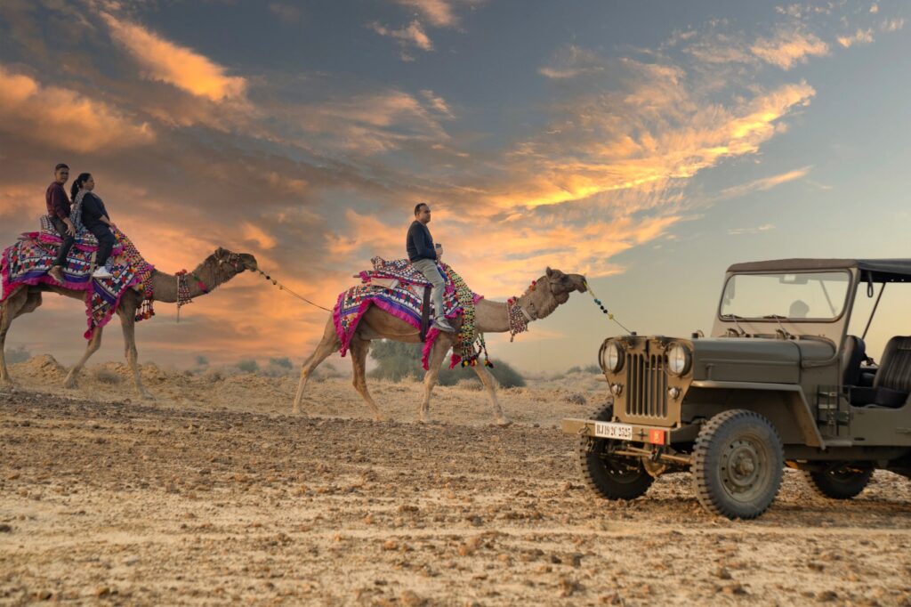 Guests enjoying camel safari beside vintage jeep during Jaisalmer desert sunset adventure.